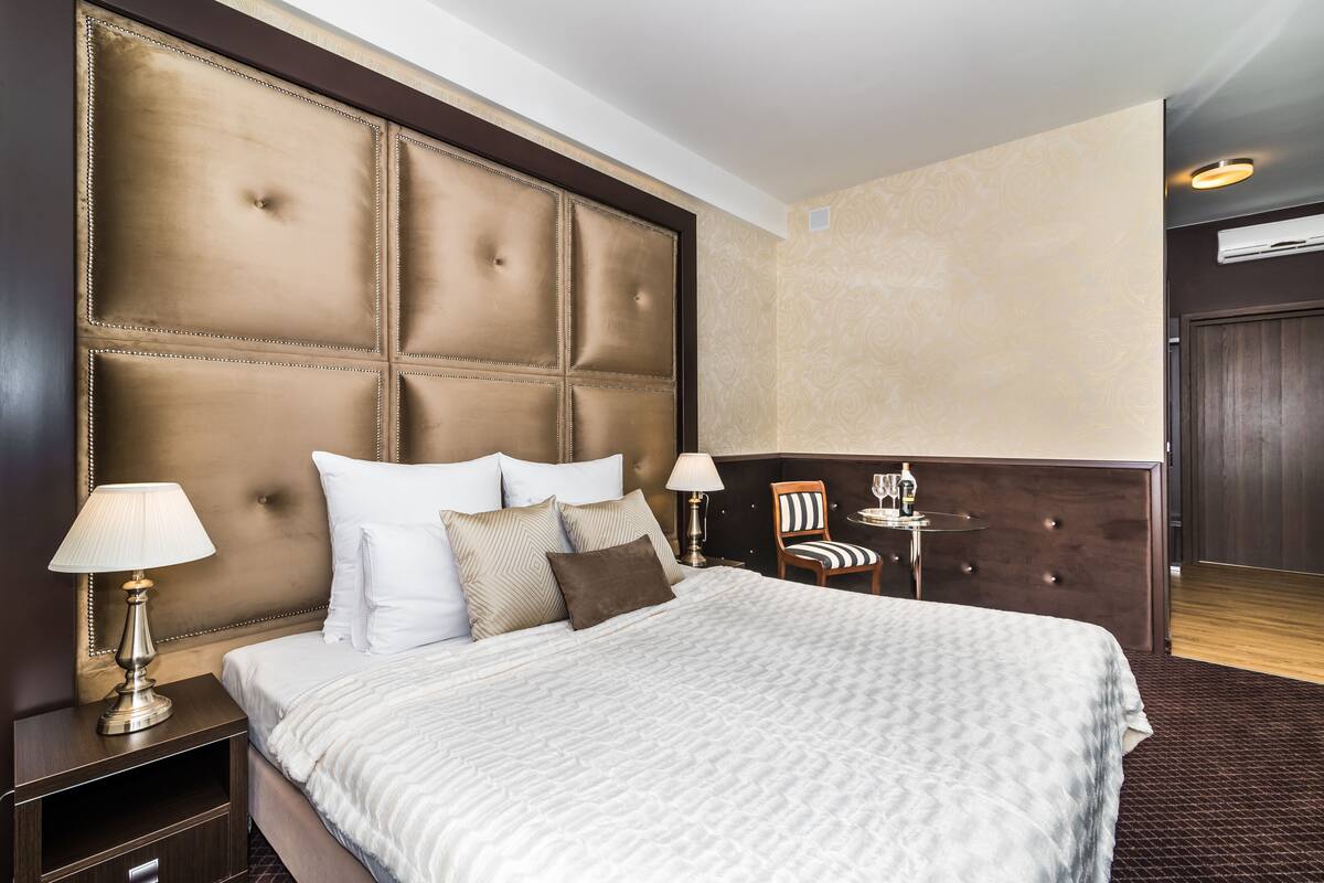 A hotel room with a large tufted headboard, a bed with white linens, two bedside tables with lamps, and a small table with a chair.
