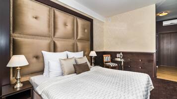 Une chambre d’hôtel dotée d’une grande tête de lit capitonnée, d’un lit avec du linge de lit blanc, de deux tables de chevet avec des lampes et d’une petite table avec une chaise.