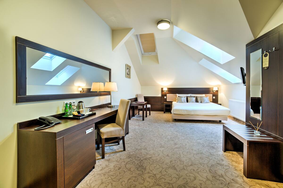 A hotel room with a large bed, a desk with a telephone, a chair, a desk lamp, a bedside table with a lamp, and a large mirror.