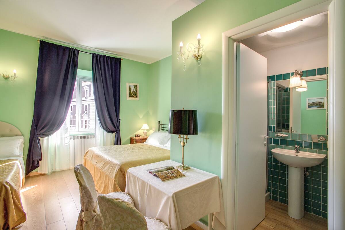 A room with two beds, a table with a book, a lamp, a chair, and a bathroom with a sink and mirror.