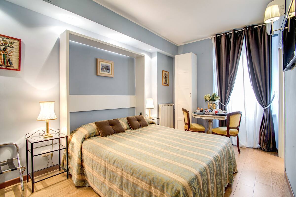 A hotel room with a large bed, a desk with a chair, a small table with a vase, and a television on the wall.
