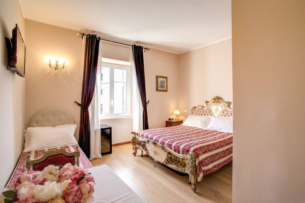 A hotel room with two beds, a chandelier, a window with curtains, and a bedside table with a lamp.