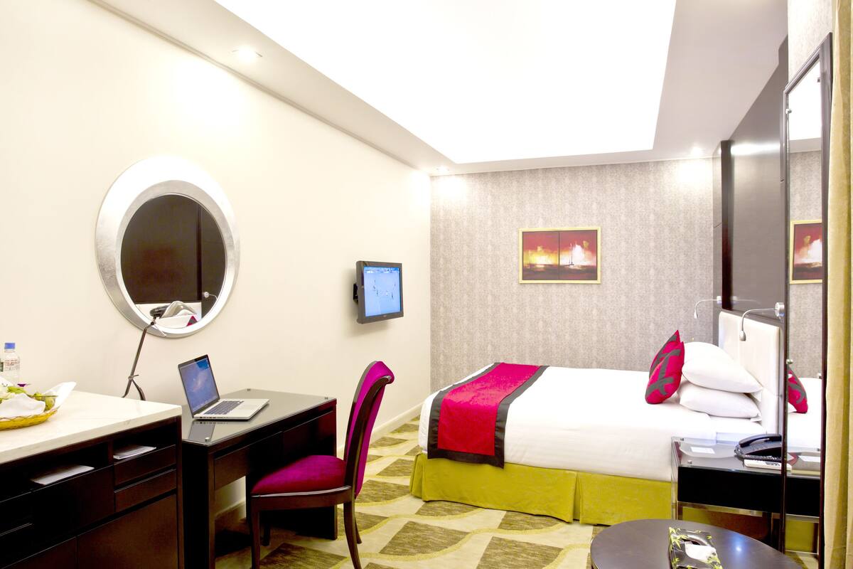 A hotel room with a bed, a desk with a laptop, a chair, a bedside table with a phone, a mirror, and a TV on the wall.