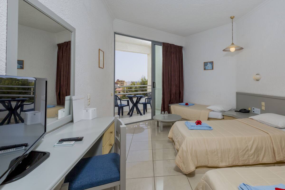 A hotel room with a bed, a desk with a computer, a chair, a table with chairs, a window with curtains, and a view of buildings outside.