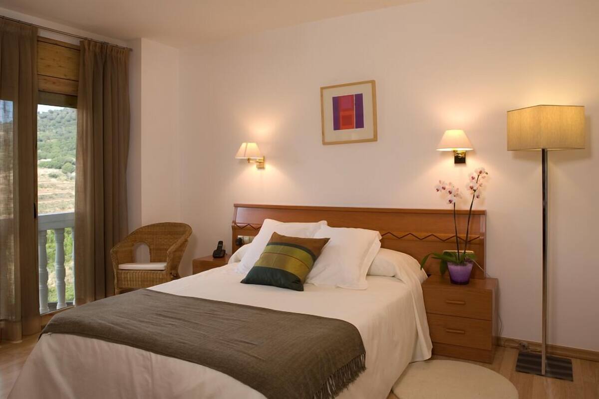 A hotel room with a bed, bedside lamps, a nightstand, a chair, a potted plant, and a view of the outdoors.