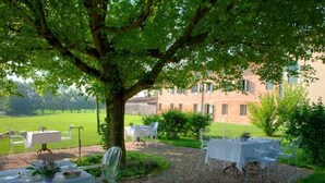 An outdoor dining area with tables set under a large tree, surrounded by a well-maintained lawn and a building in the background. - Relais Corte Guastalla (Sona)