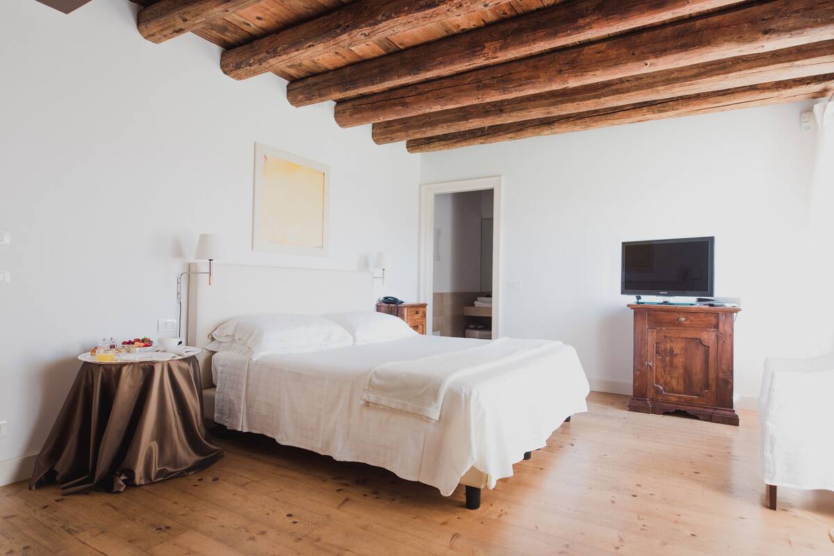 A bedroom with a wooden ceiling, a bed with white bedding, a bedside table with a lamp, a television on a wooden cabinet, and a door leading to another room.