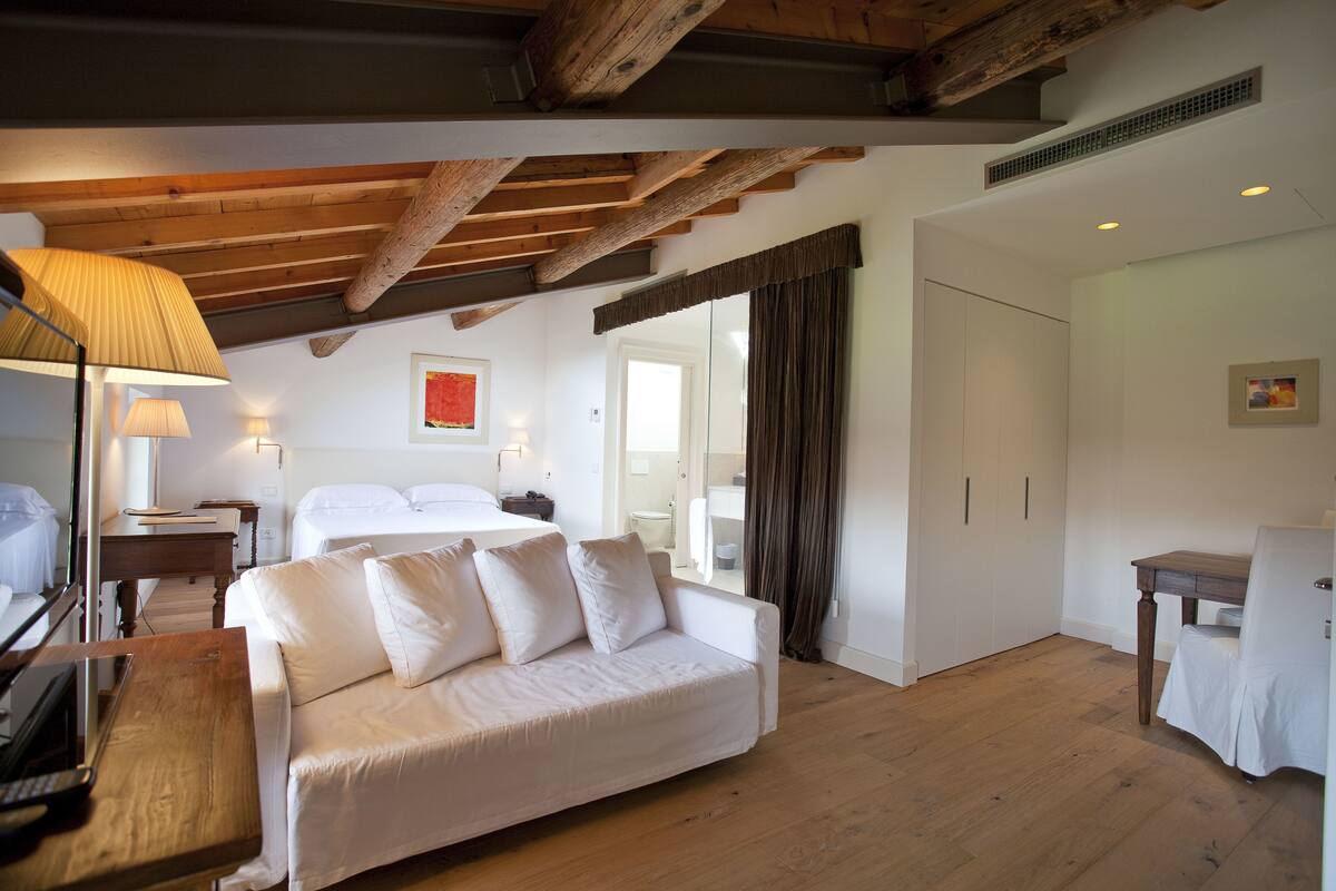 A hotel room with a wooden ceiling, a white sofa, a bedside table, a lamp, a bed, and a door leading to a bathroom.