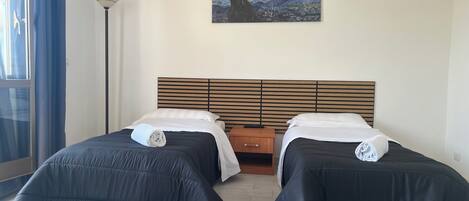 A hotel room with two beds, a wooden headboard, a painting of a night sky, a floor lamp, and a window with blue curtains.