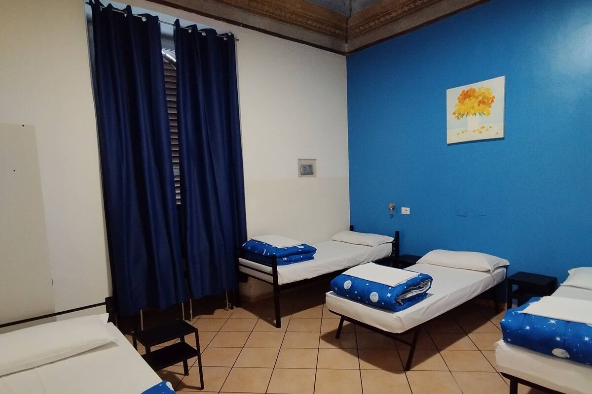A room with four single beds, each with a blue and white cover, a blue curtain, and a small table beside each bed. The room has a tiled floor and a blue wall with a painting.