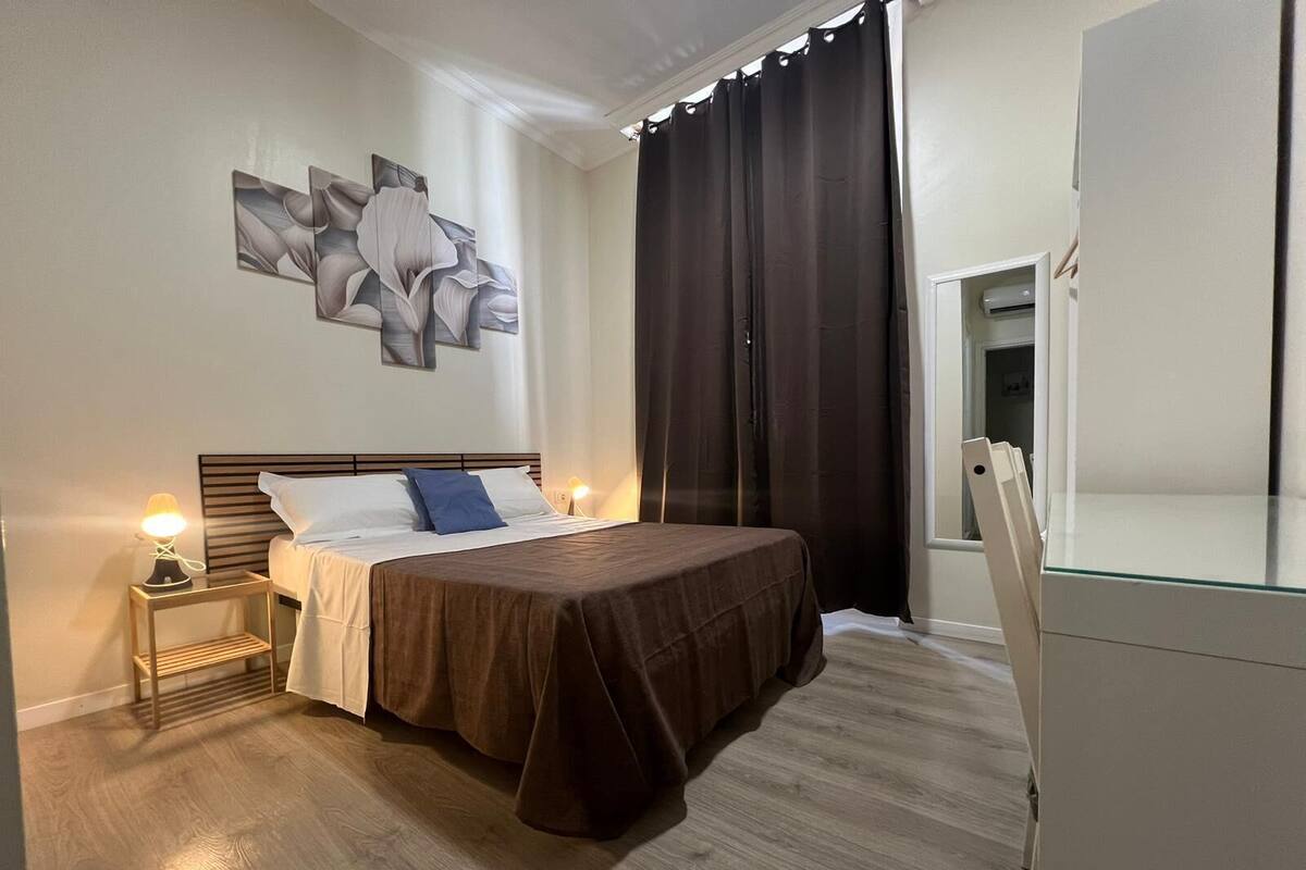 A modern hotel room with a large bed, a nightstand with a lamp, a wall-mounted artwork, and a large window with curtains.