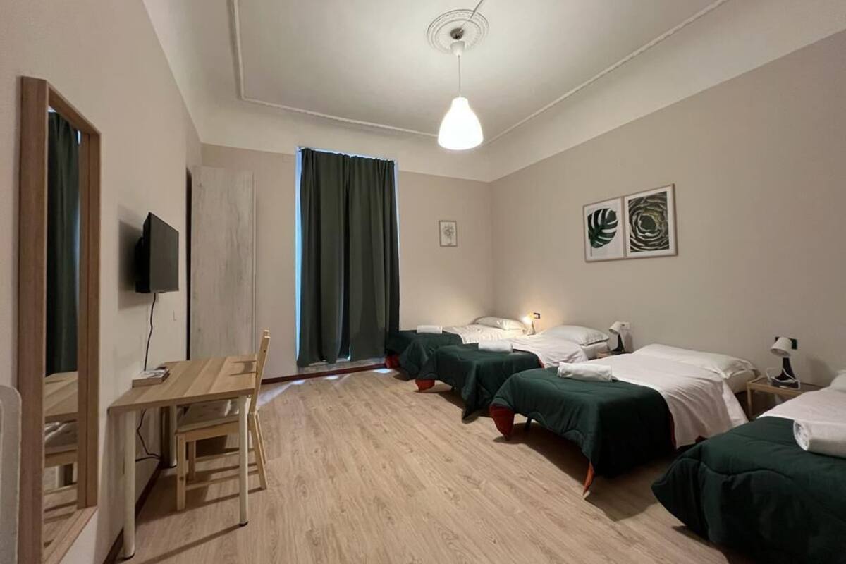 A hotel room with a wooden desk, two beds with green bedding, a ceiling light, and artwork on the wall.
