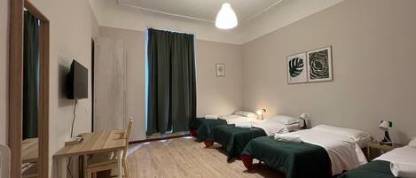 A hotel room with a wooden desk, two beds with green bedding, a ceiling light, and artwork on the wall.