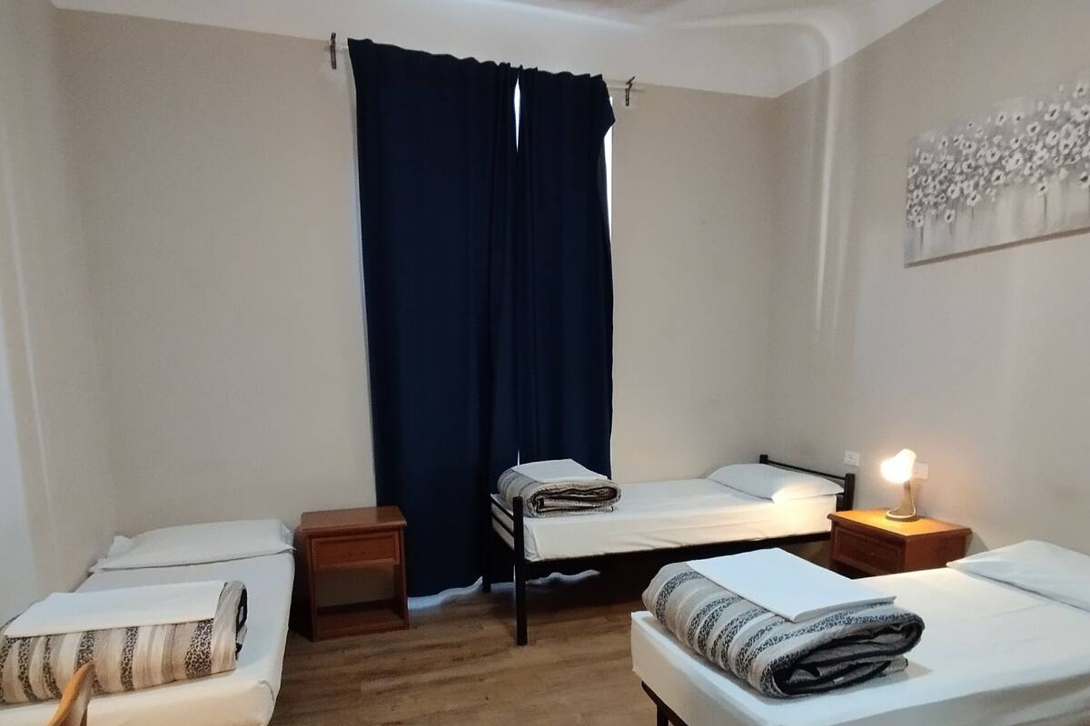 A room with two single beds, a wooden floor, a nightstand with a lamp, and a window with dark curtains.