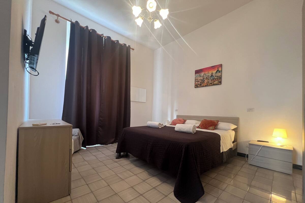A hotel room with a bed, a bedside table, a nightstand, a lamp, a painting on the wall, and a window with curtains.