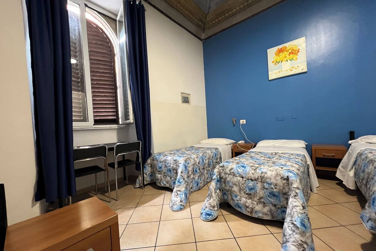 A room with two beds, a desk, and a chair. There is a window with blinds and curtains.