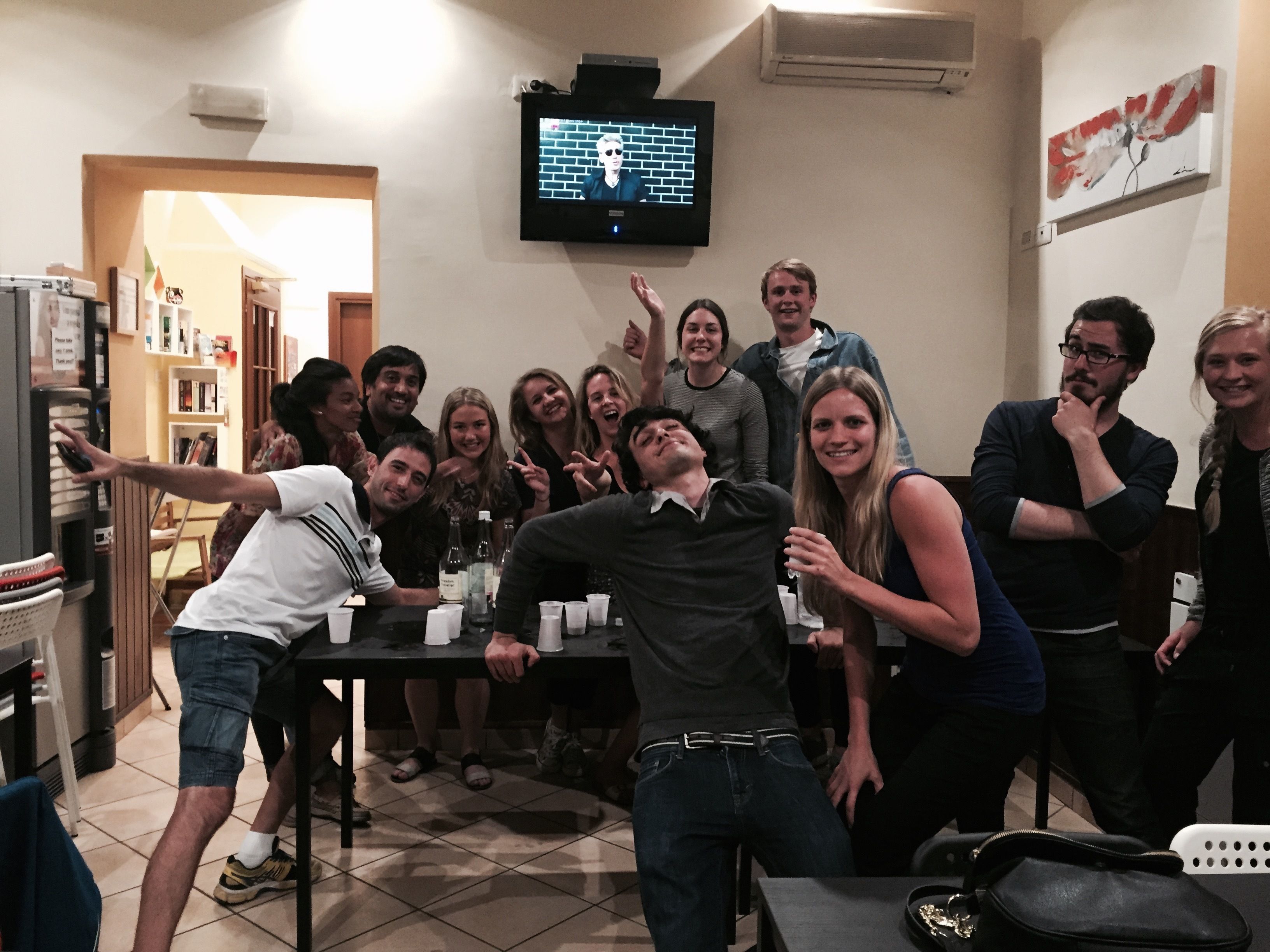A group of people in a room with a television mounted on the wall, a table with cups and bottles, and a refrigerator.