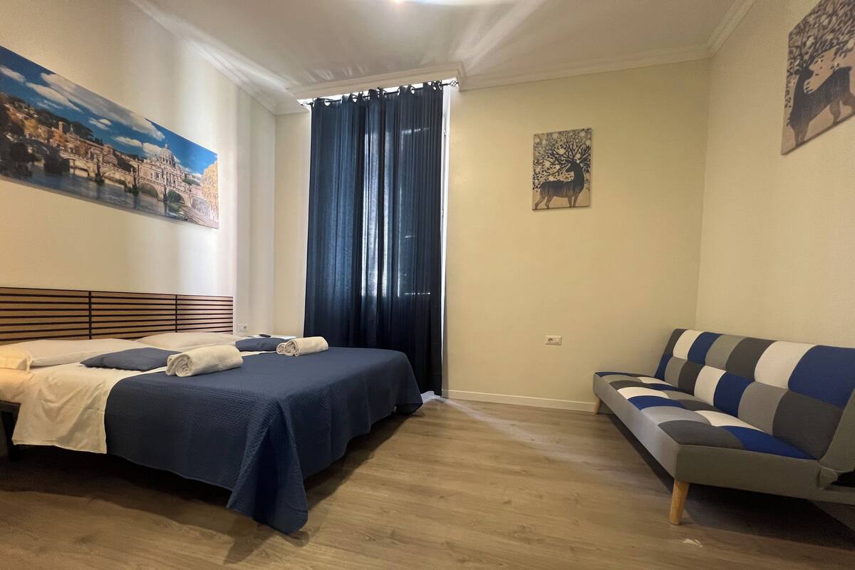 A bedroom with two beds, a sofa, a large window with curtains, and a painting on the wall.
