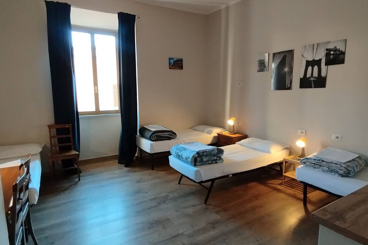 A room with two beds, a desk, and a chair. There is a window with curtains, a small table with a lamp, and a wall with pictures.