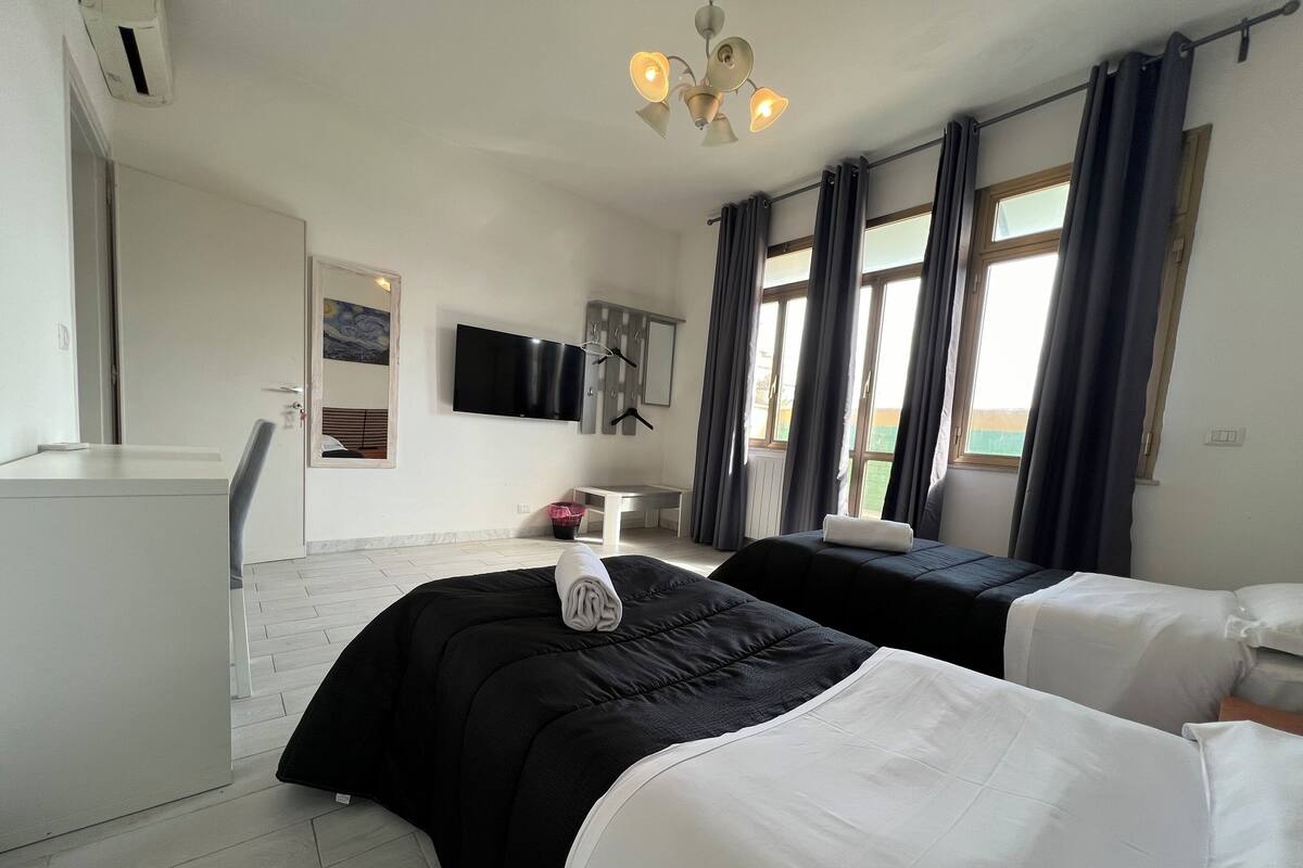 A hotel room with two beds, a desk, a chair, a TV, and a window with curtains.