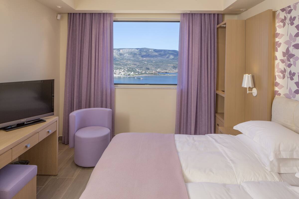A hotel room with a bed, a TV, a desk, a chair, and a view of a body of water and mountains.