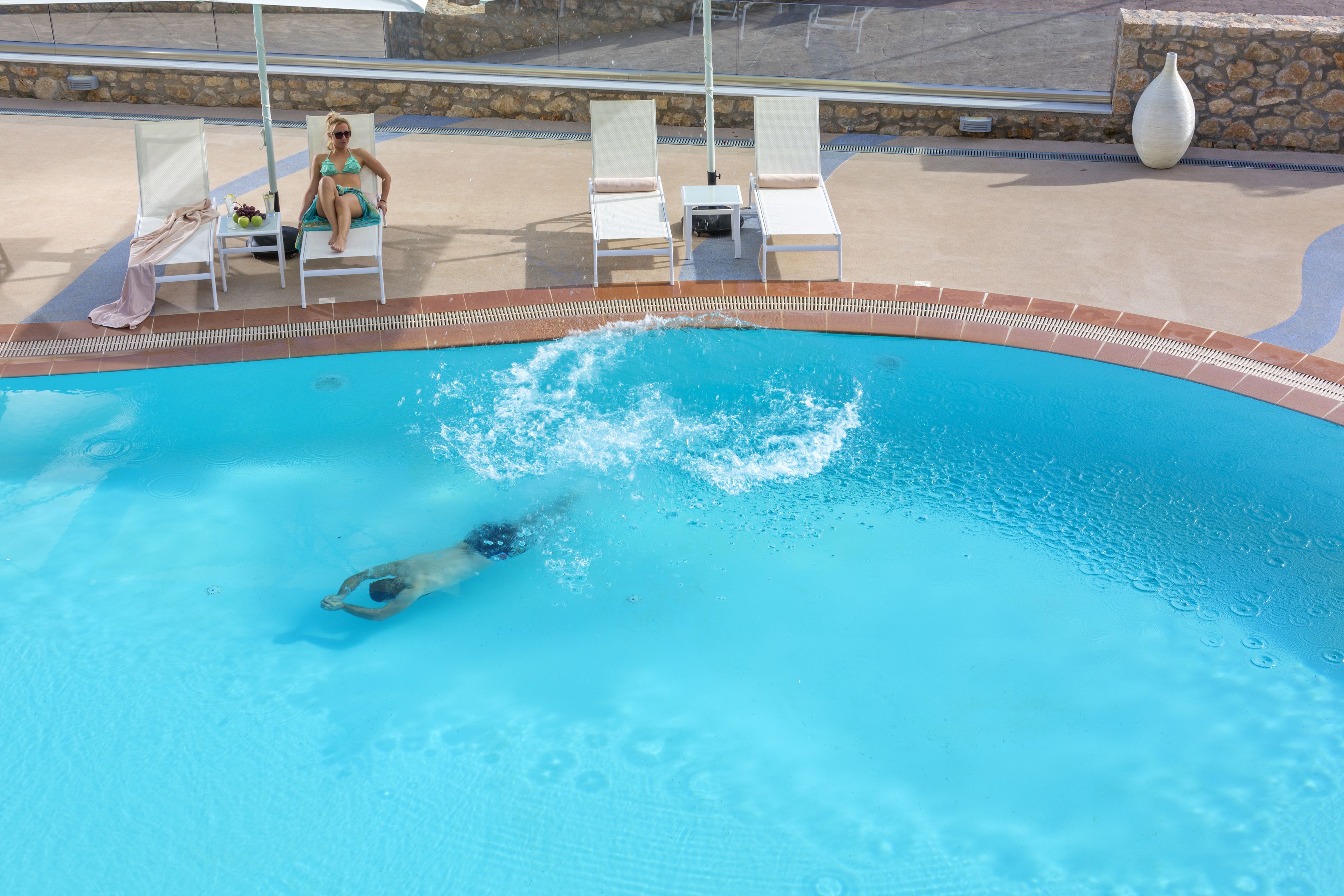 A swimming pool with a person swimming, lounge chairs, and a stone wall in the background.