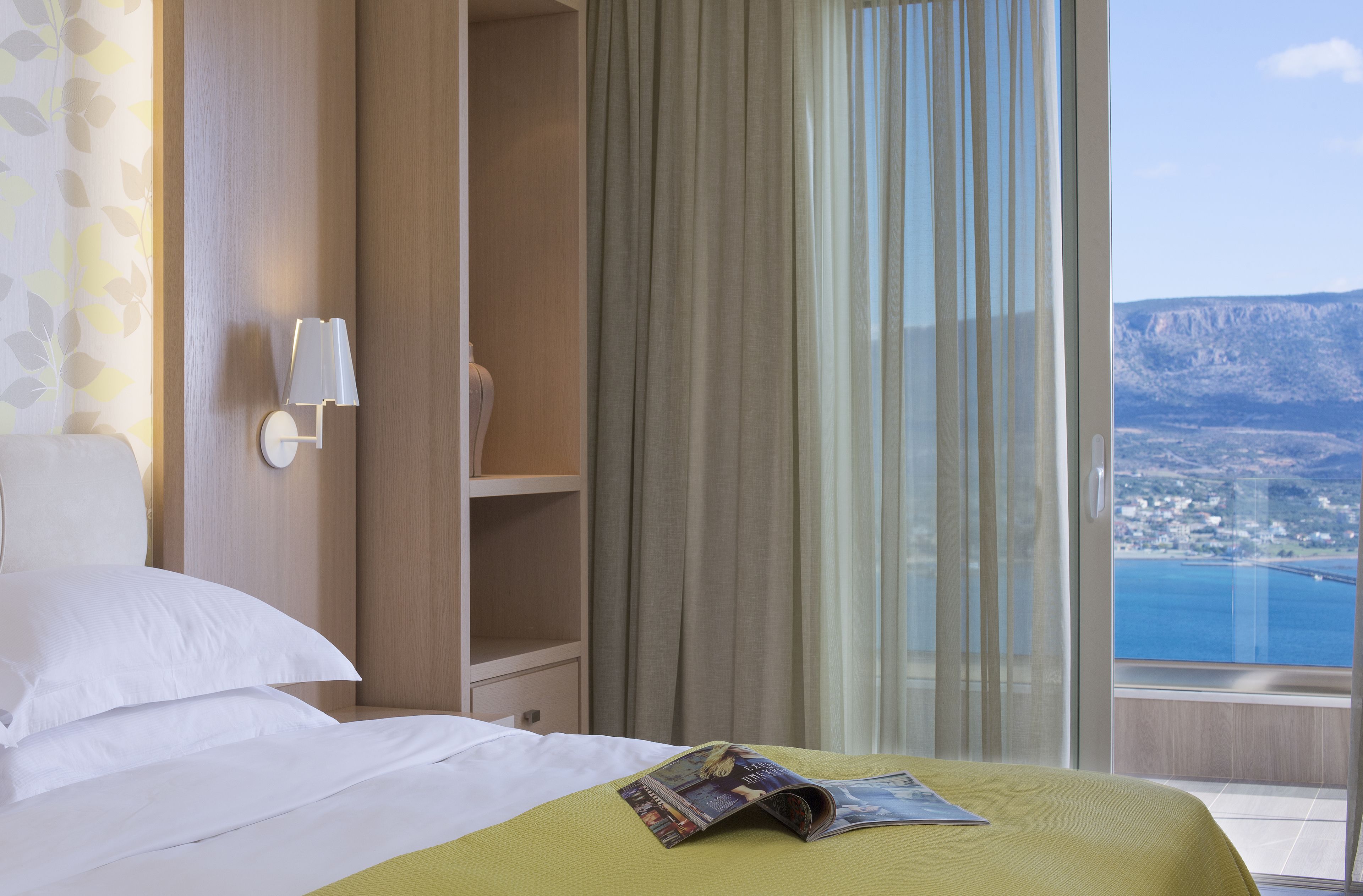 A hotel room with a bed, a nightstand, a lamp, a window with curtains, and a view of a coastal landscape.