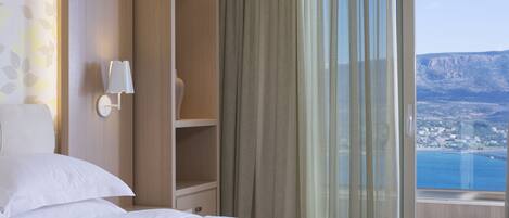 A hotel room with a bed, a nightstand, a lamp, a window with curtains, and a view of a coastal landscape.