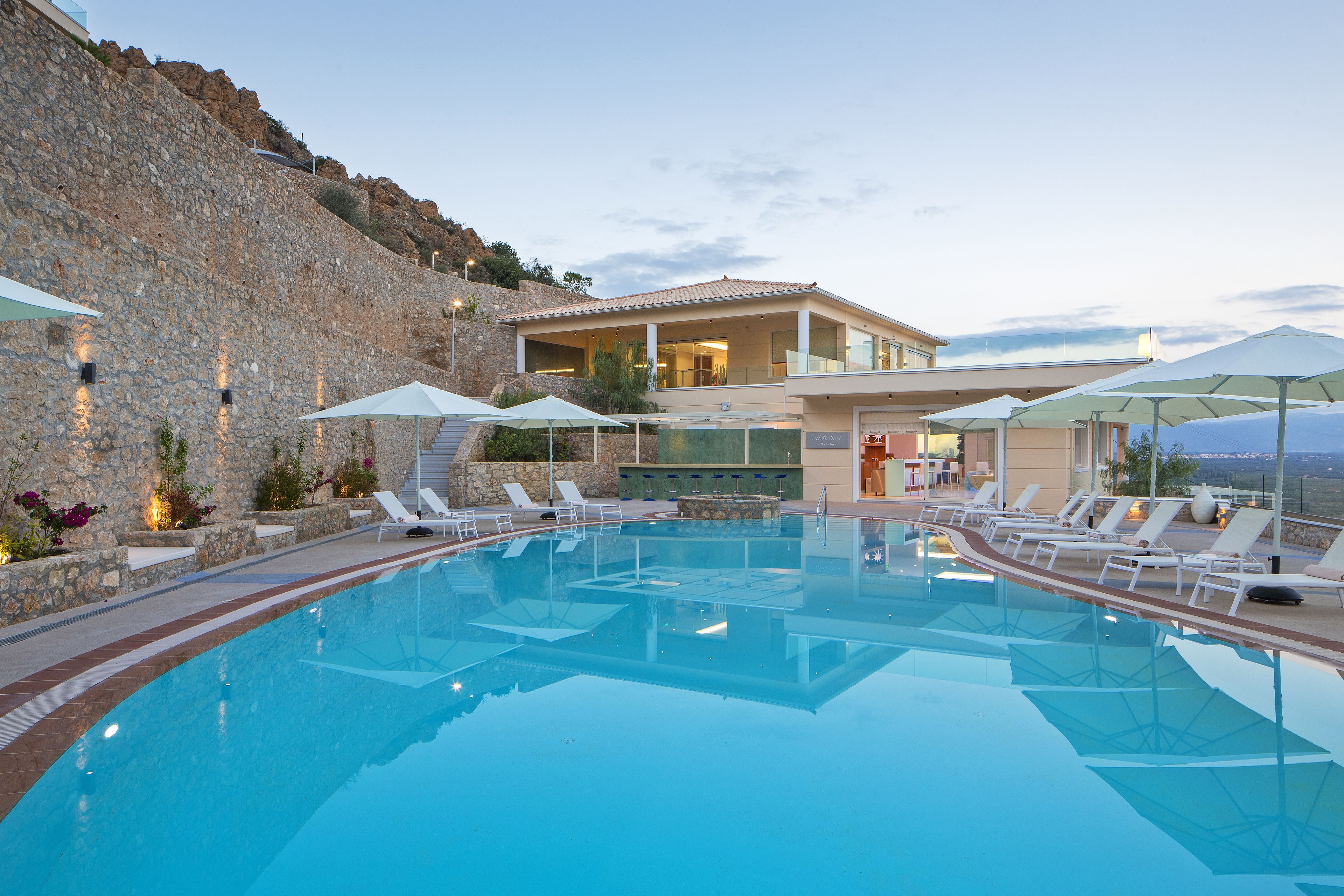 A swimming pool with lounge chairs and umbrellas, a stone wall, and a building with large windows.