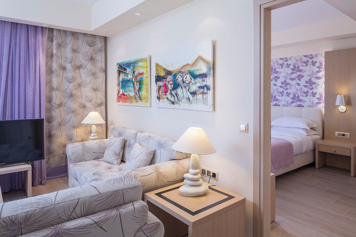 Una habitación de hotel moderna con sofá, televisión, obras de arte en las paredes y una cama con colcha morada.