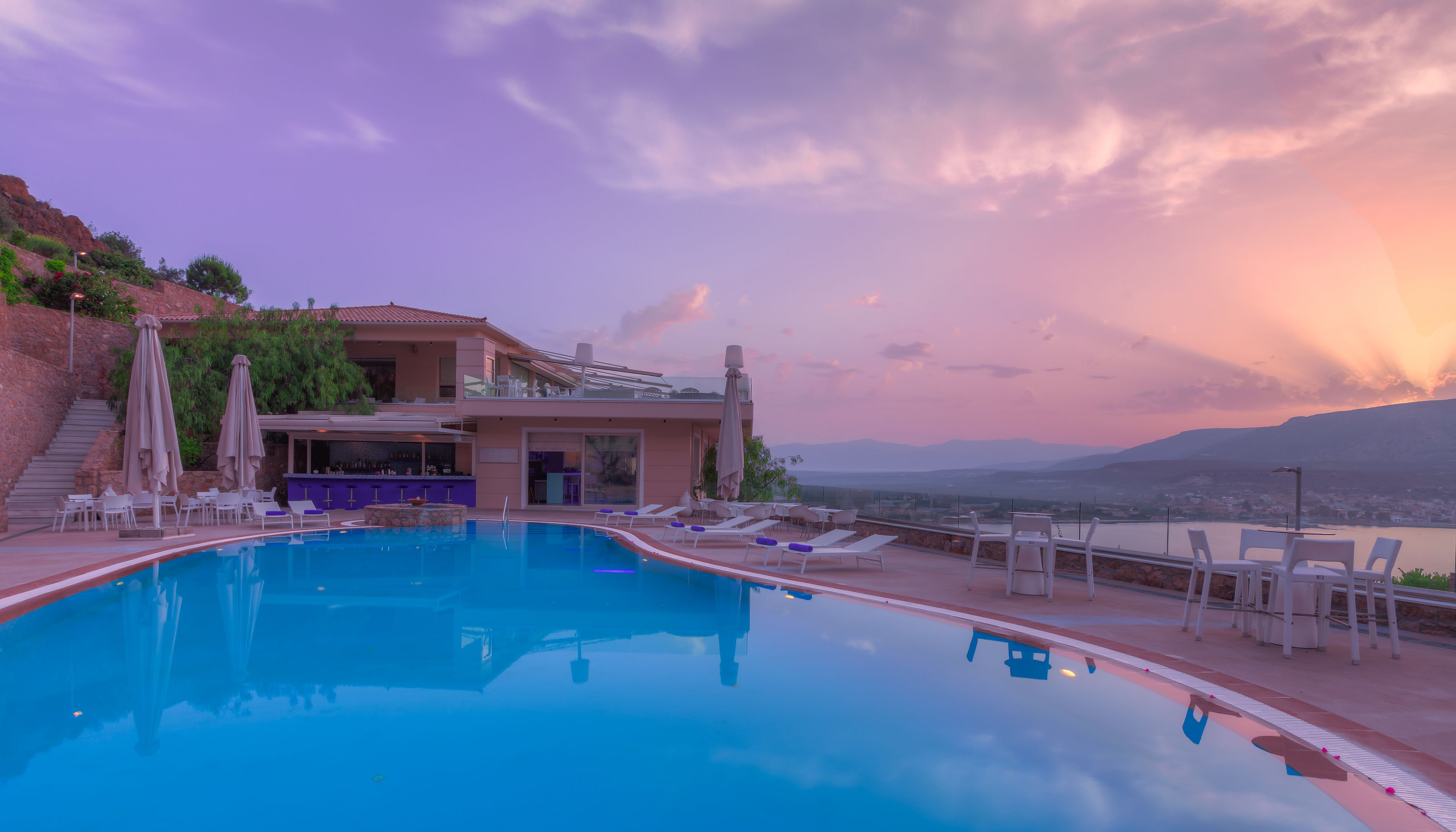 Una piscina con tumbonas y sombrillas, un área de bar y vistas a montañas y una masa de agua.