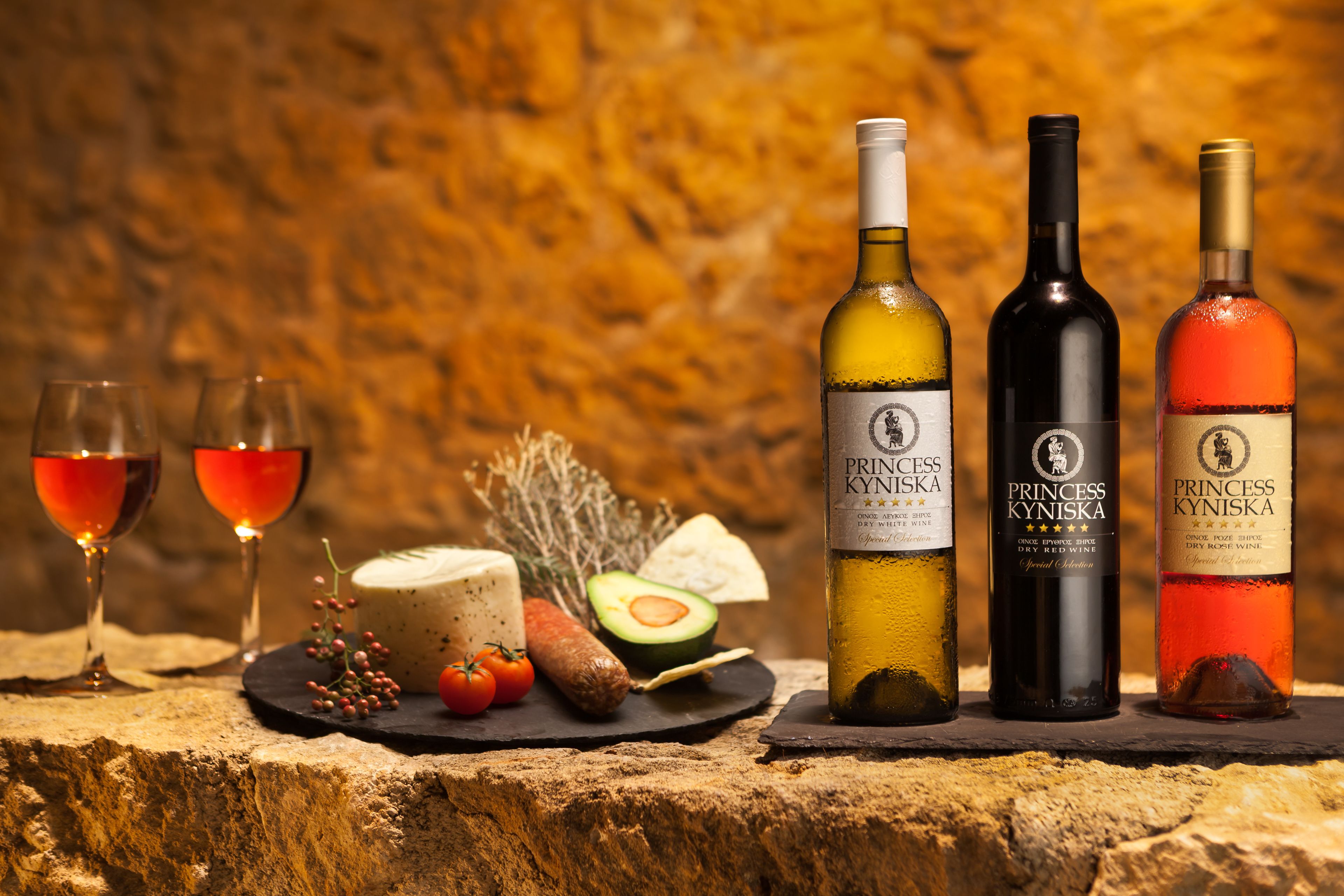 Tres botellas de vino Princess Kyniska y dos copas de vino tinto sobre una superficie de piedra, junto con alimentos.