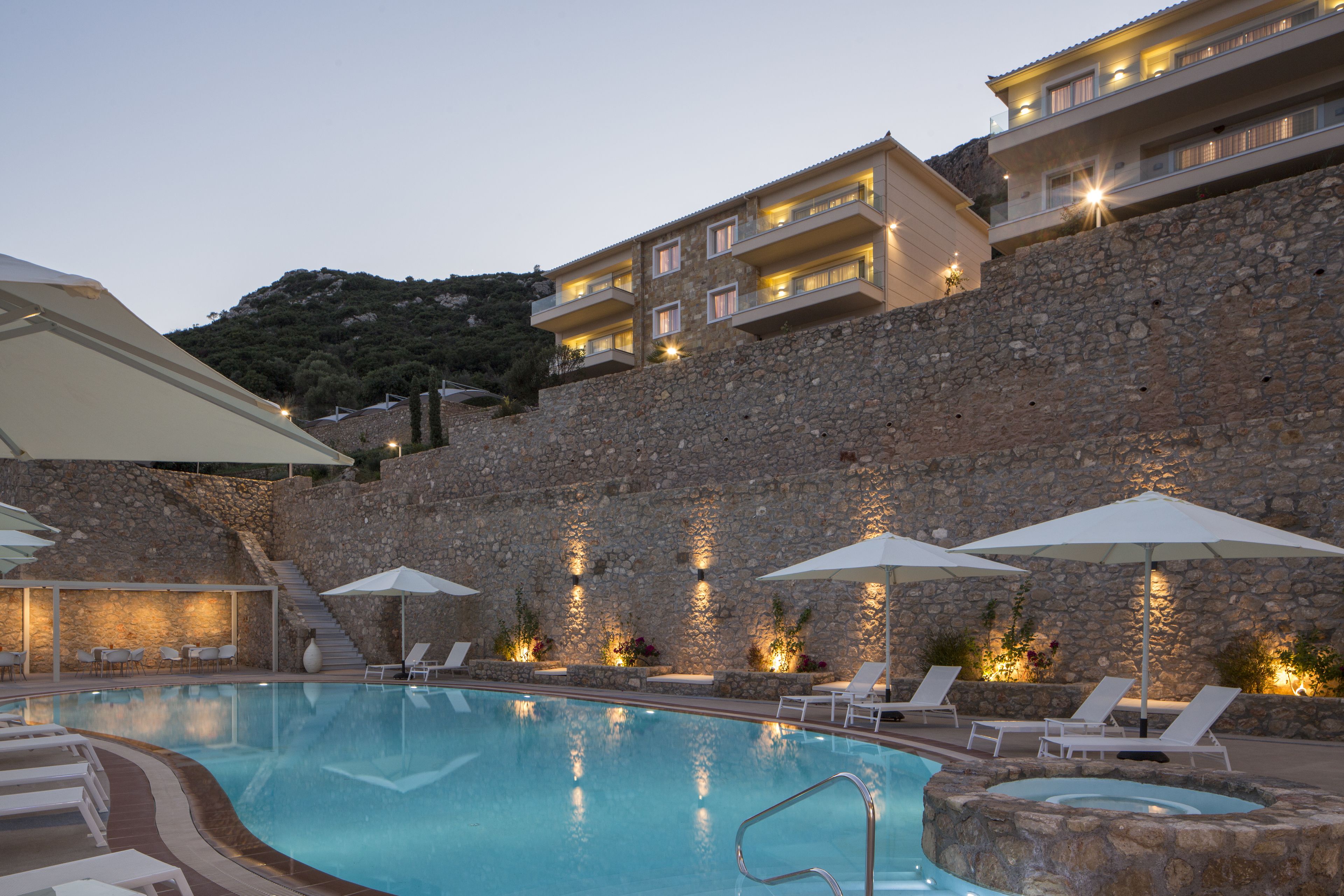 Une piscine avec des chaises longues et des parasols, un mur en pierre et un immeuble à plusieurs étages doté de balcons.