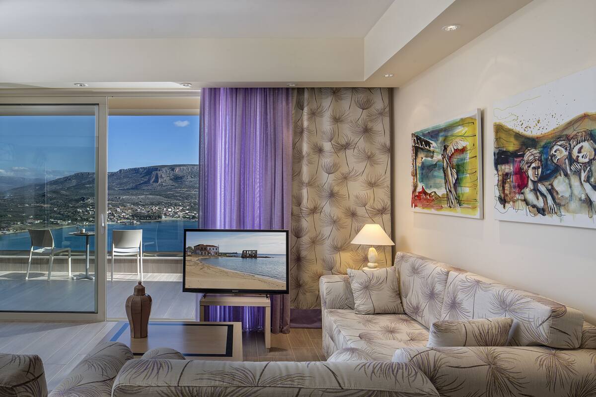 A modern living room with a large TV, a sofa, and a view of a coastal landscape through a glass door.