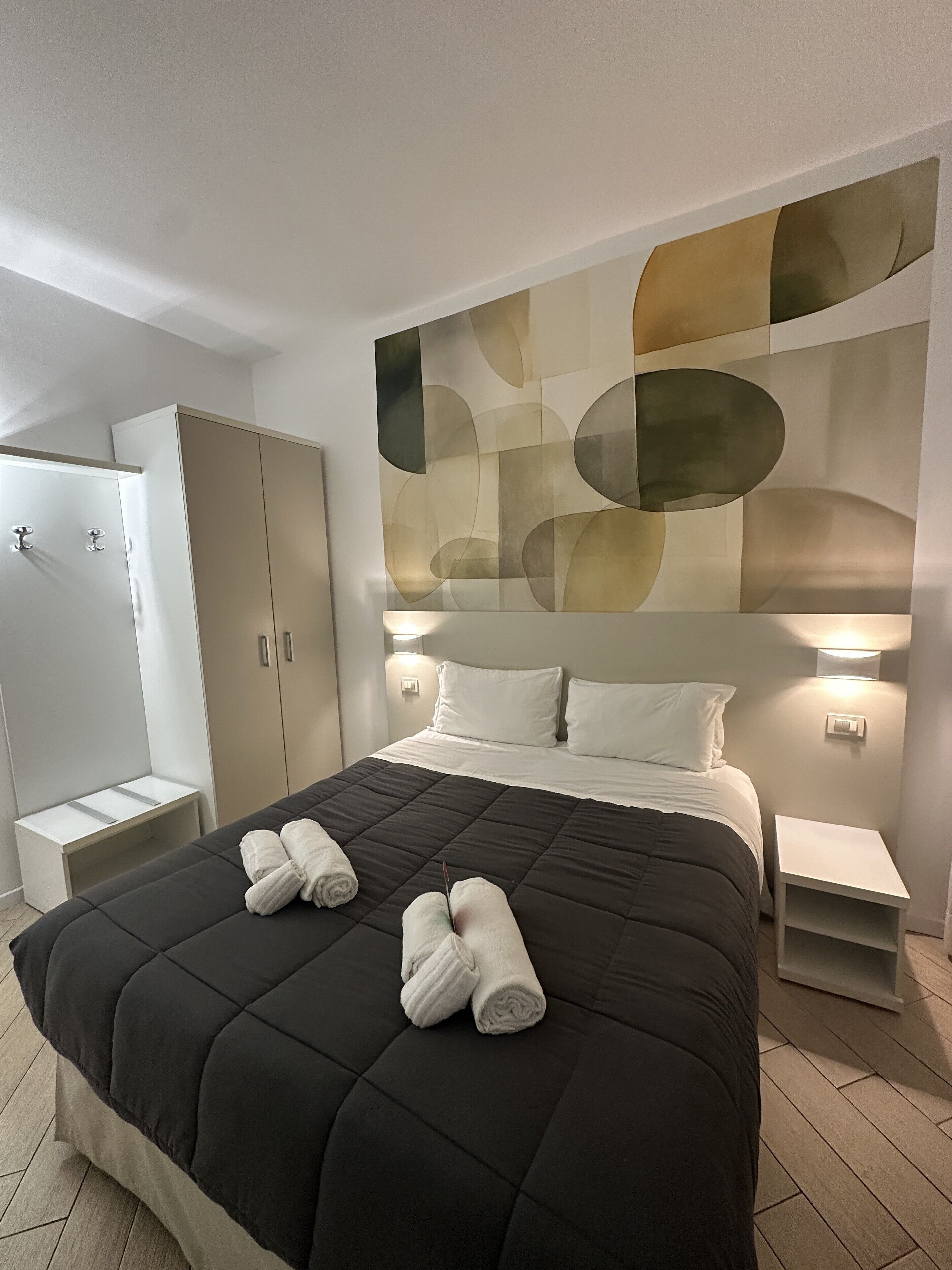 A modern hotel room with a large bed, two bedside tables, a wardrobe, and abstract wall art.