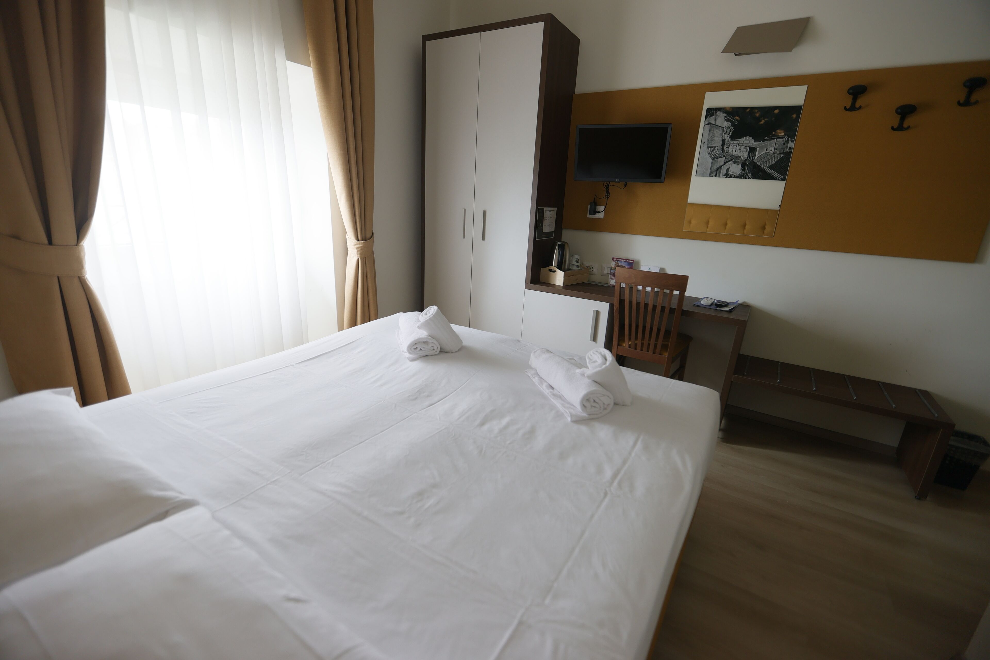 A hotel room with a large bed, a wooden desk, a chair, a television, and a bulletin board.