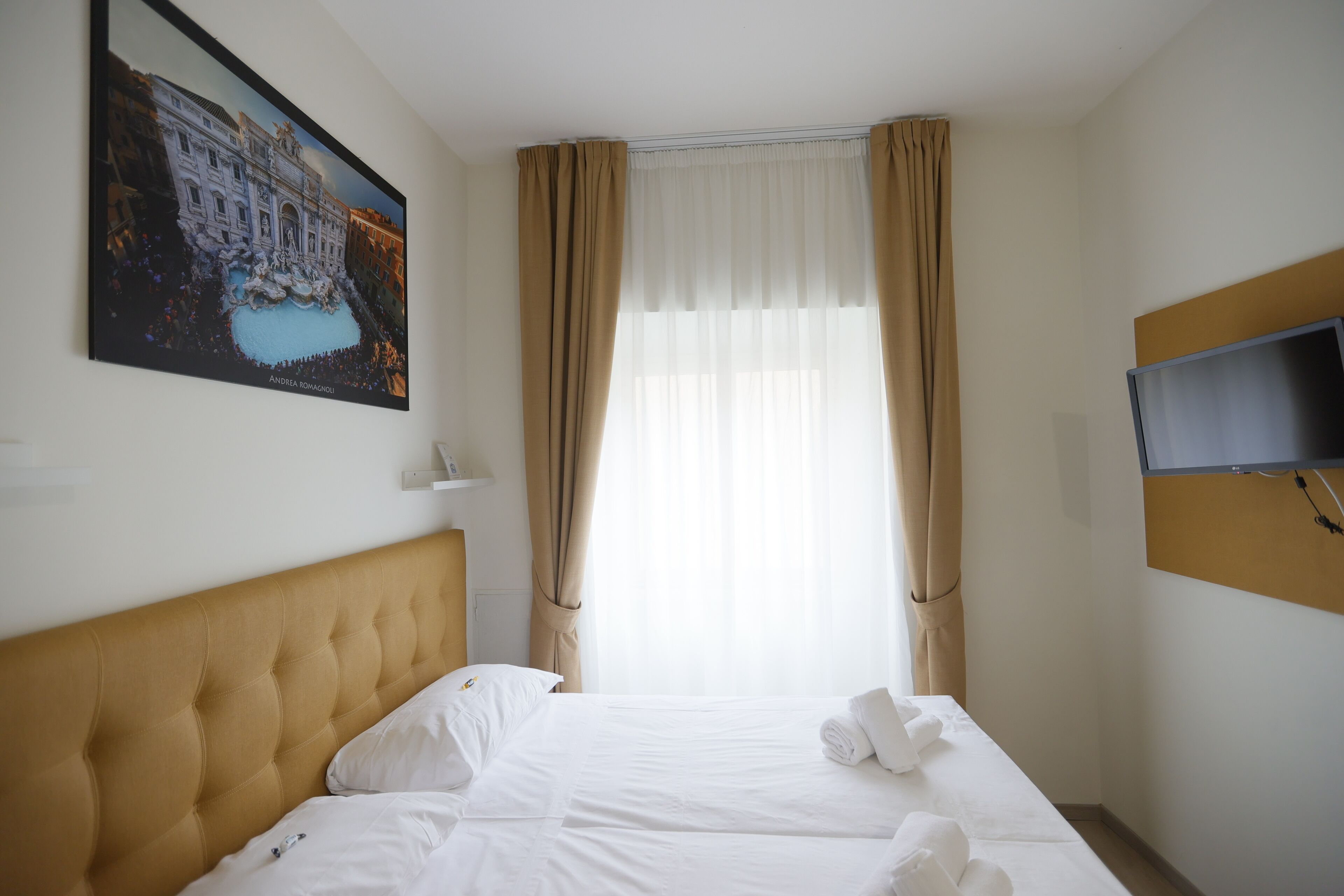 A hotel room with a bed, white linens, a headboard, a painting of a building, a TV, and curtains.