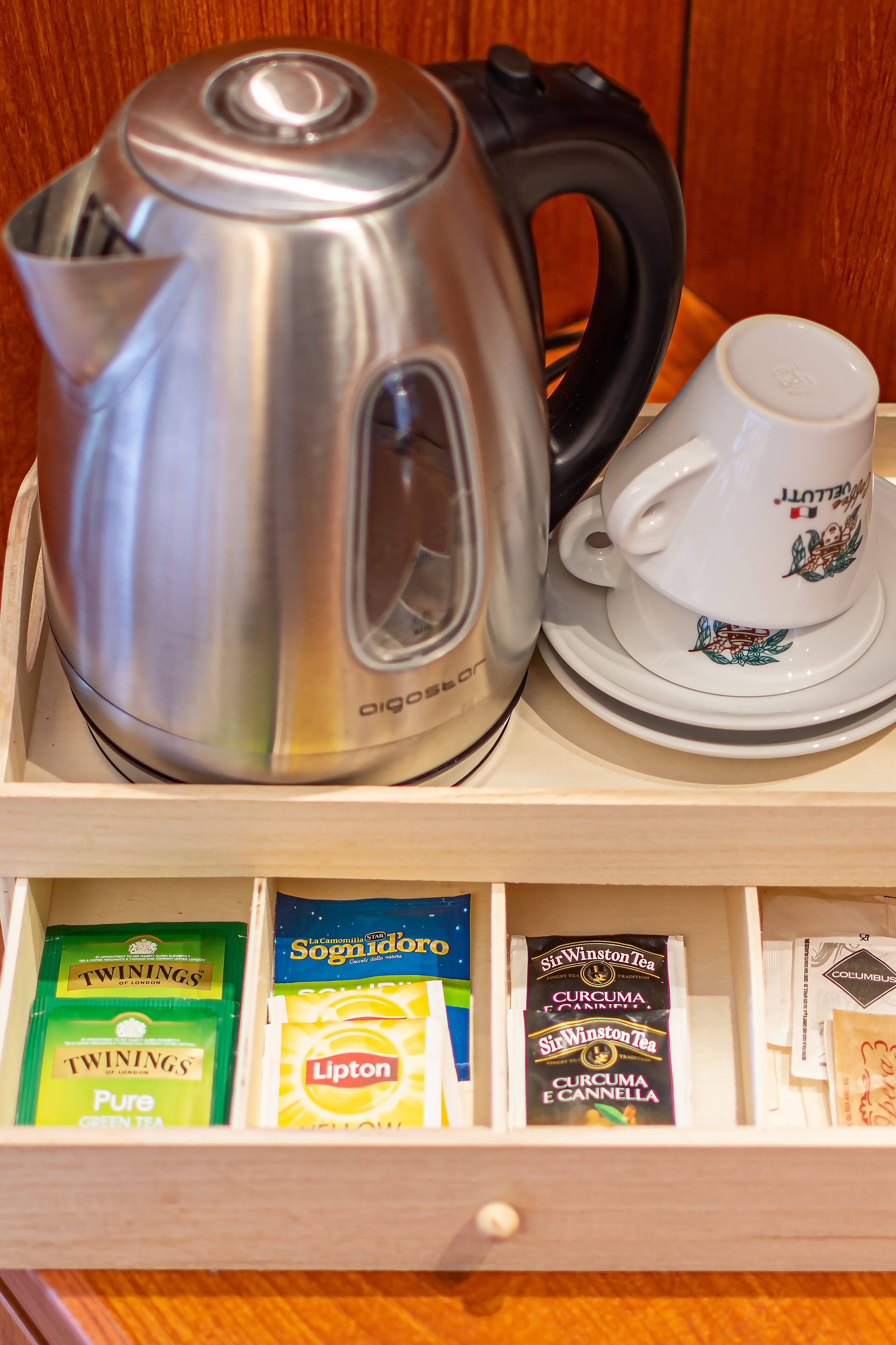 A stainless steel kettle, a white teacup with a saucer, and a drawer containing tea bags and Lipton tea boxes.