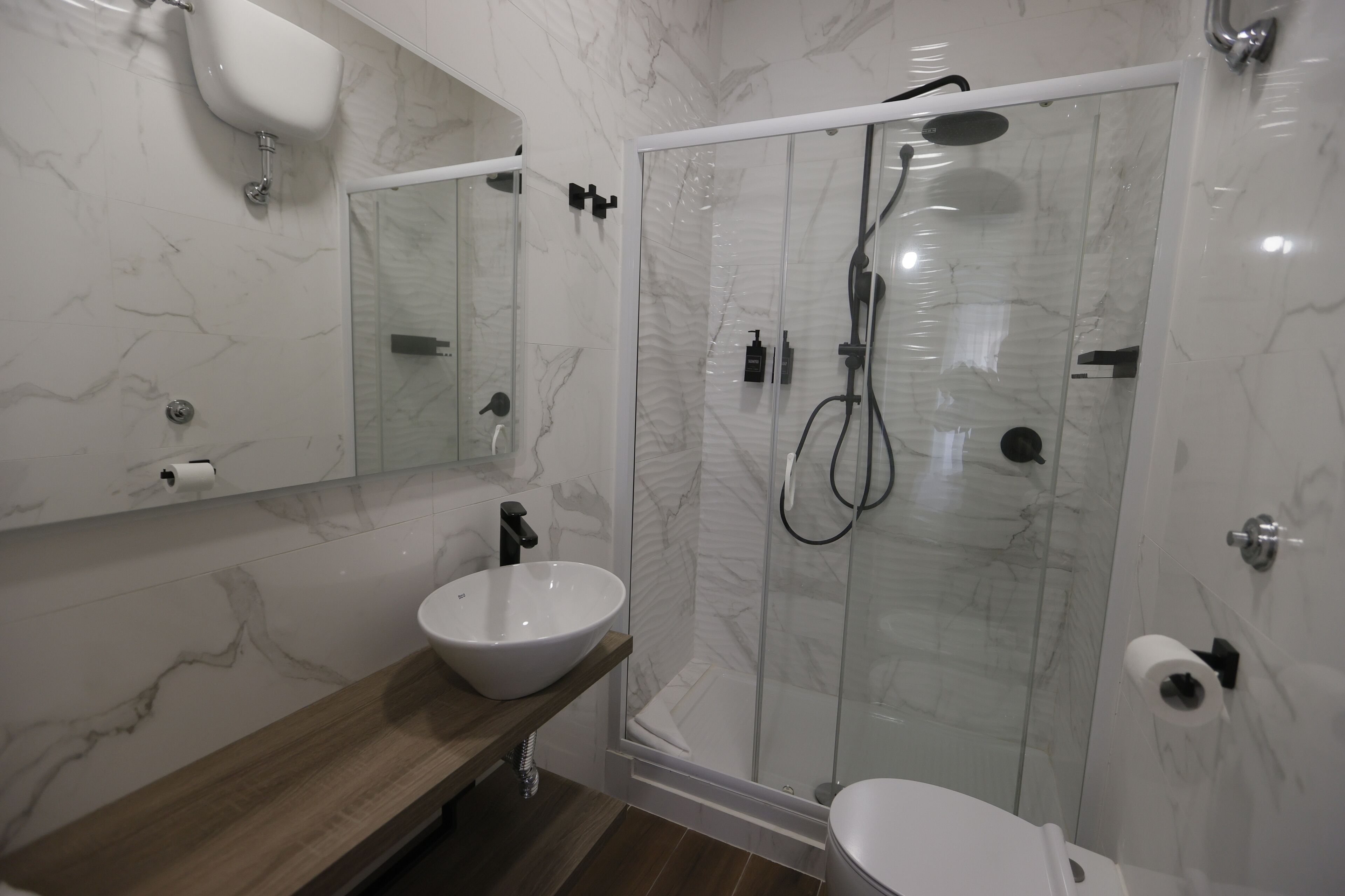 A modern bathroom with a marble backsplash, a wooden vanity, a white sink, a glass-enclosed shower with a handheld showerhead, and a toilet.