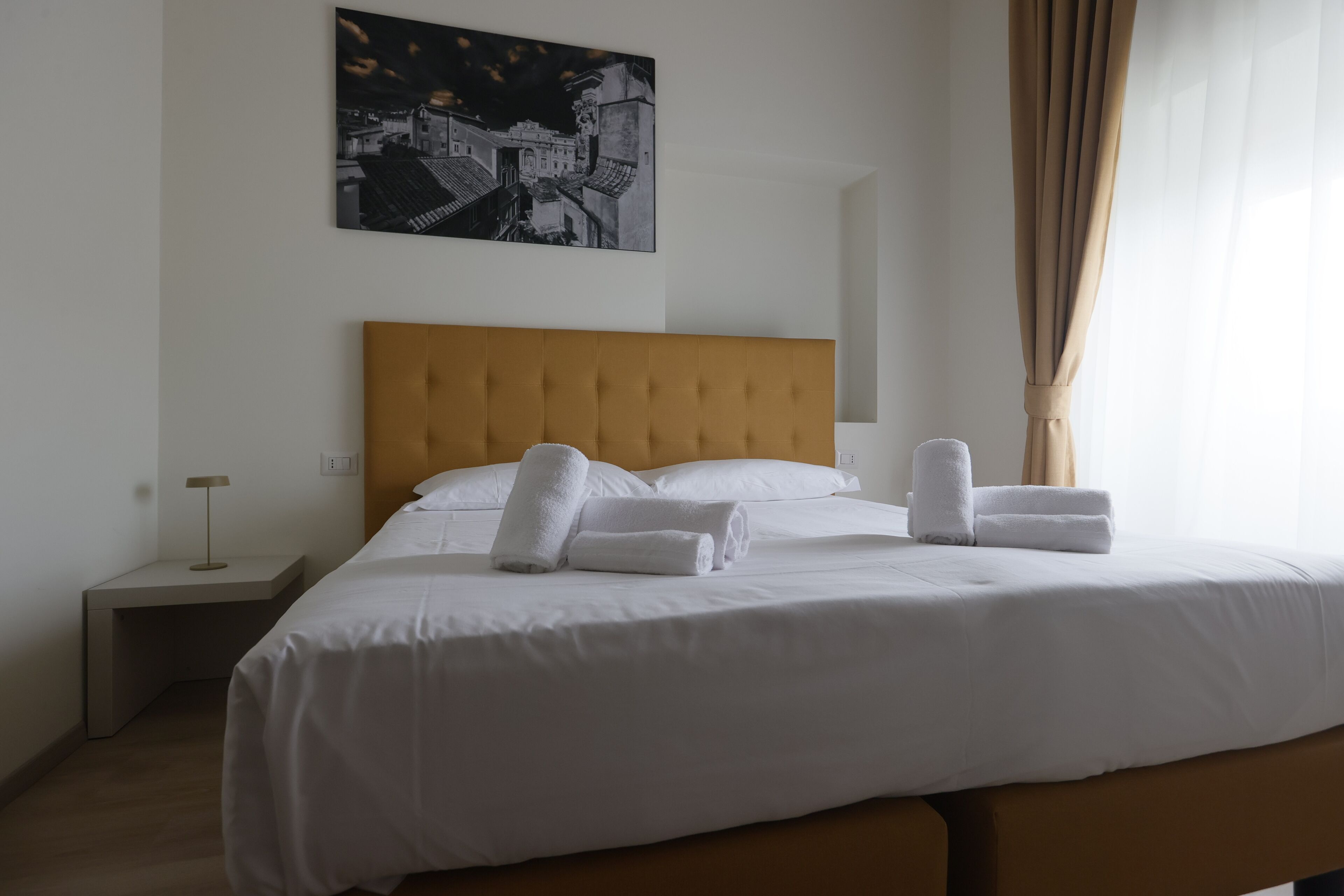 A neatly made bed with white linens and towels, a wooden headboard, a bedside table with a lamp, and a cityscape painting on the wall.