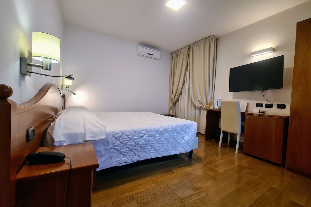 A hotel room with a bed, a desk, a chair, a television, and an air conditioning unit.