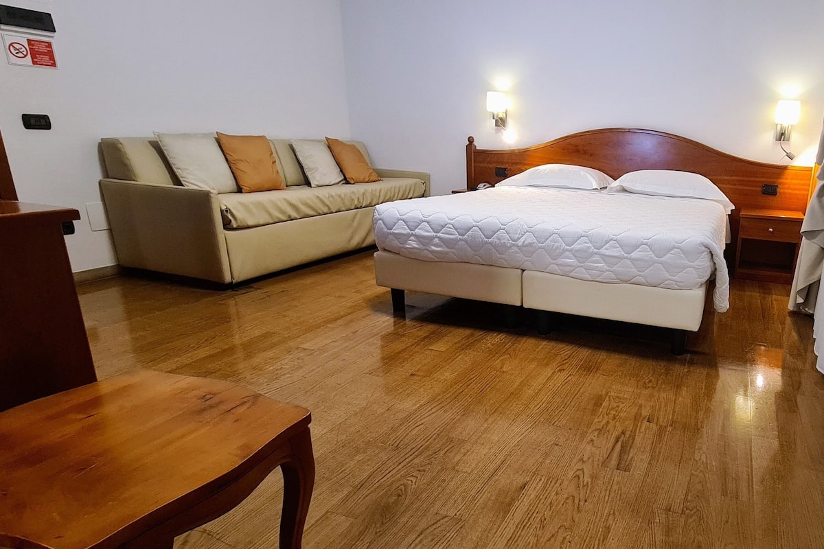 A hotel room with a bed, a sofa, a wooden nightstand, and a wooden chair.