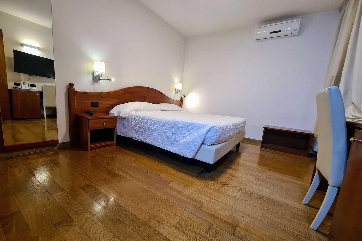 A bedroom with a wooden bed, bedside lamps, a flat-screen TV, and an air conditioning unit.