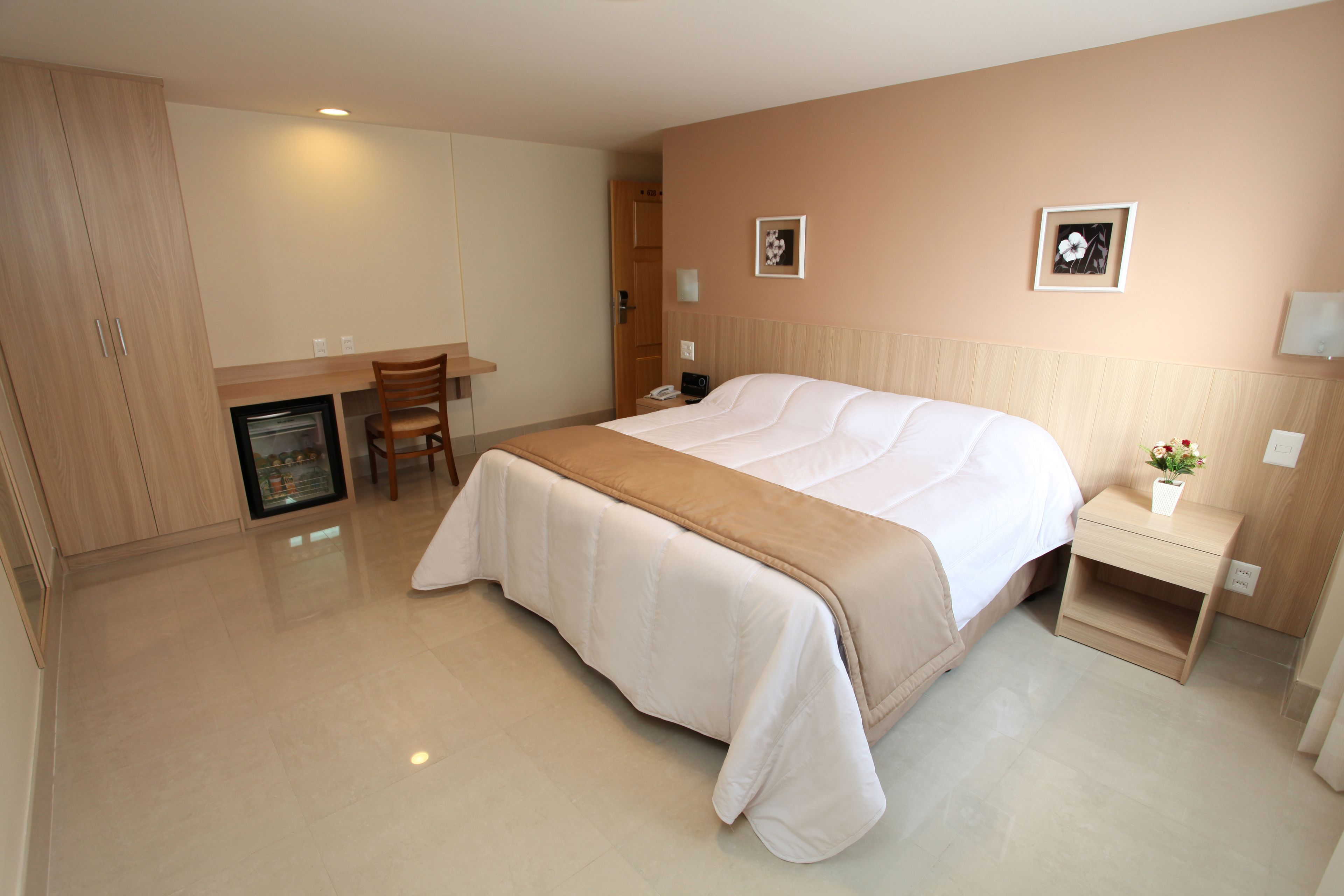 A hotel room with a bed, bedside table, chair, desk, and a small refrigerator.