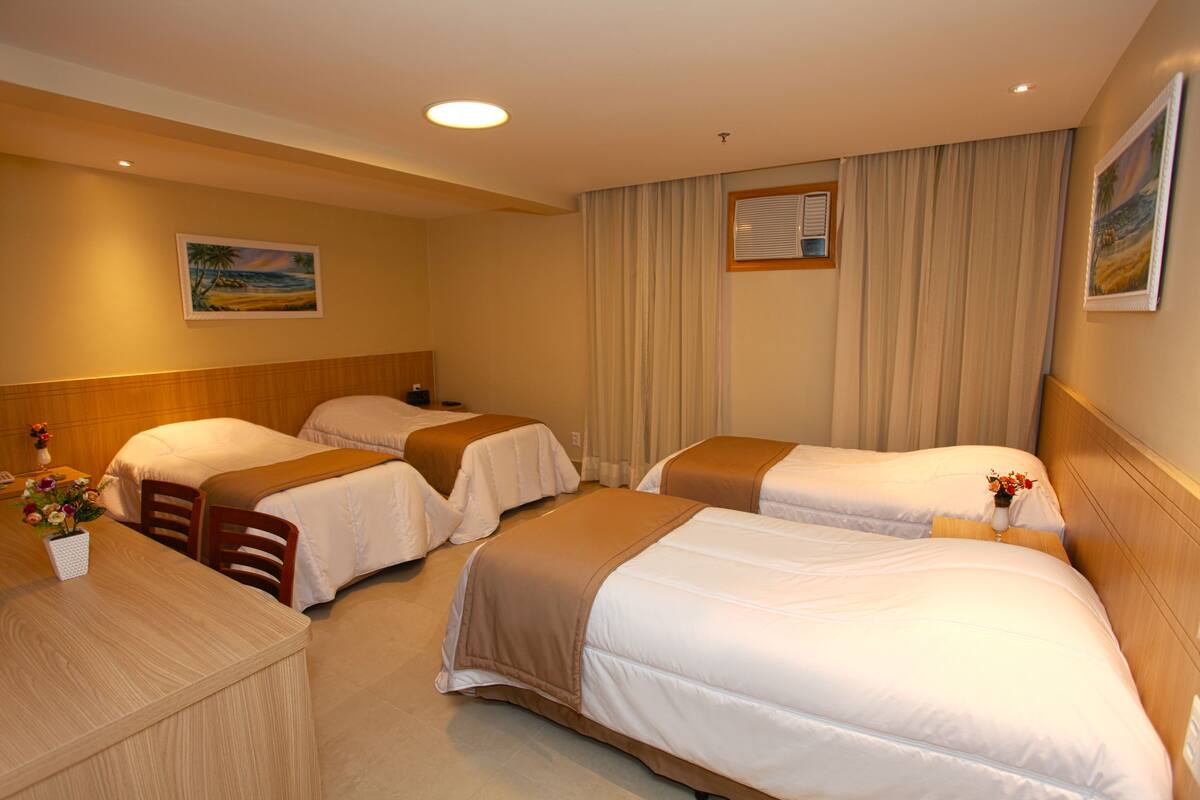 A hotel room with three beds, a wooden headboard, a desk with a vase of flowers, and a painting on the wall.