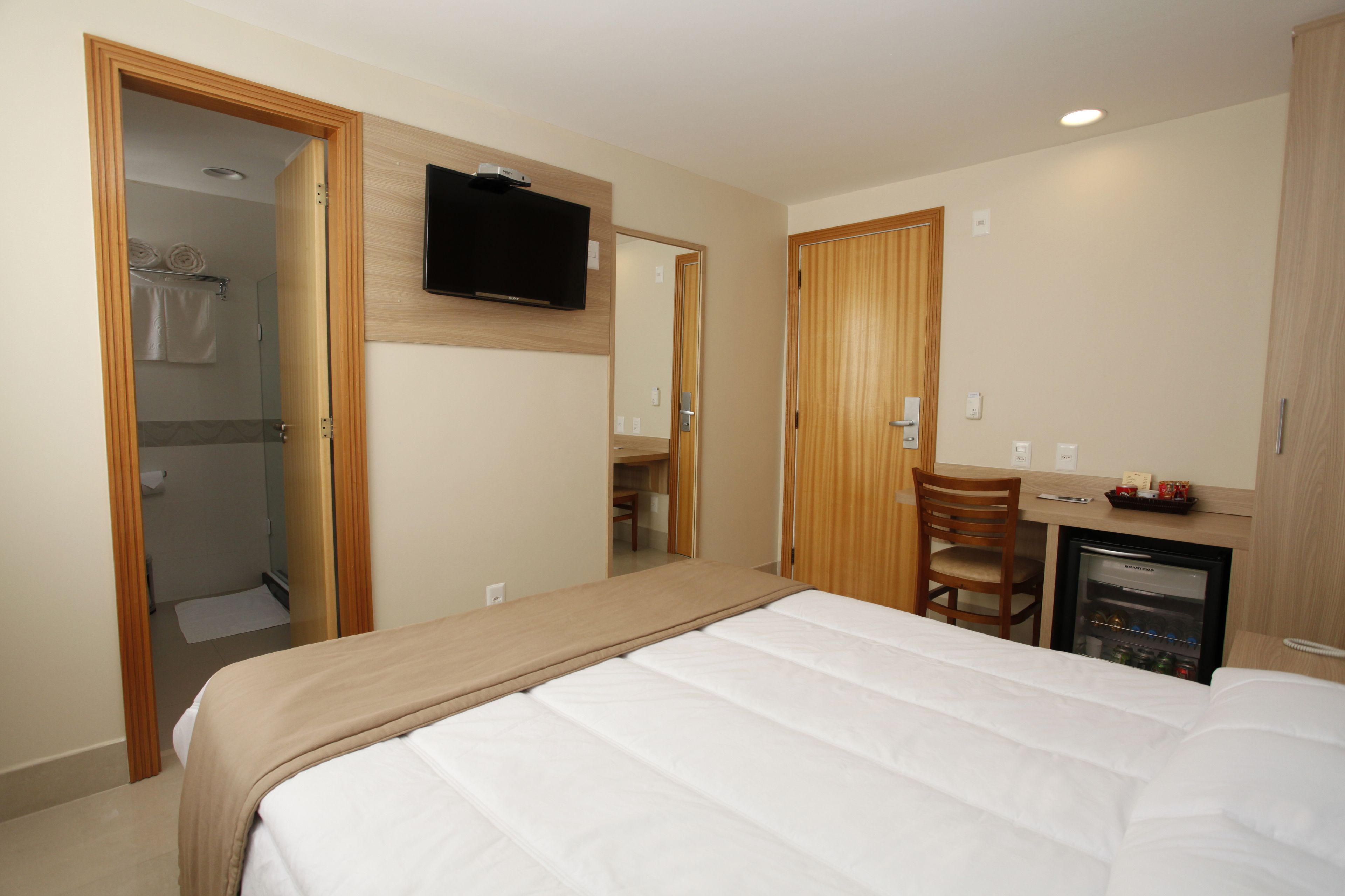 Quarto de hotel com cama, televisão, escrivaninha com cadeira, mini-geladeira e chuveiro.