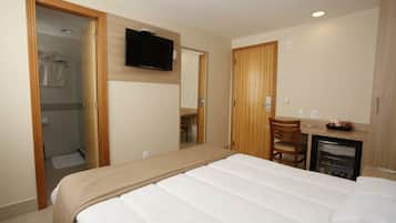Quarto de hotel com cama, televisão, escrivaninha com cadeira, mini-geladeira e chuveiro.