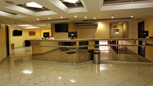 A hotel lobby with a reception desk, a flat-screen TV, an air conditioning unit, and a trash can. - Elegance Praia Hotel (Rio de Janeiro)