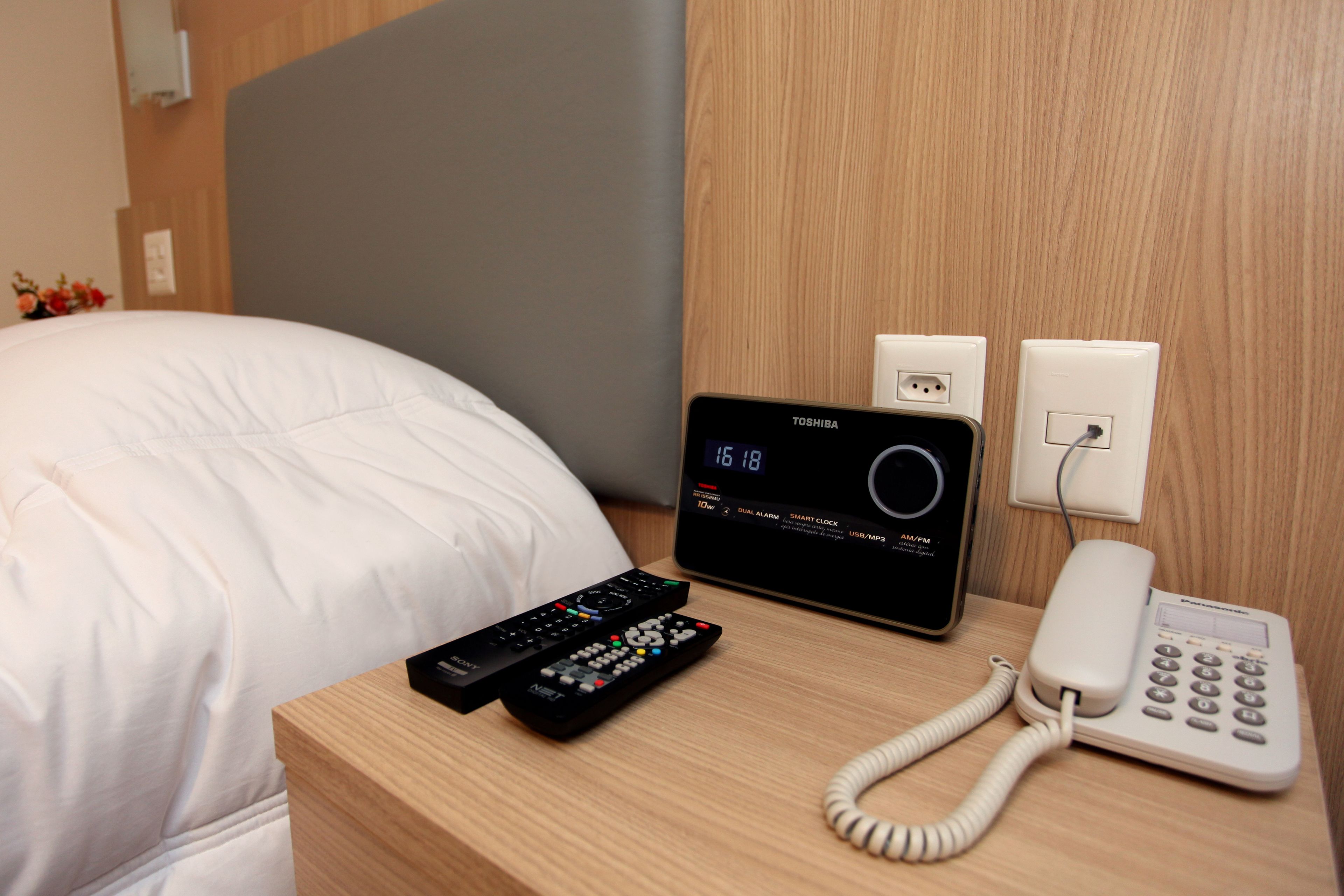A hotel room with a bed, a bedside table with a digital clock, a telephone, and two remote controls.