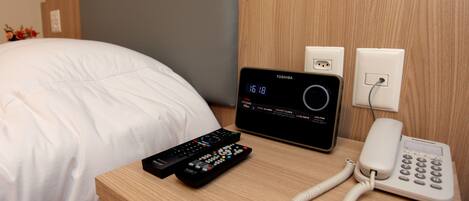 A hotel room with a bed, a bedside table with a digital clock, a telephone, and two remote controls.